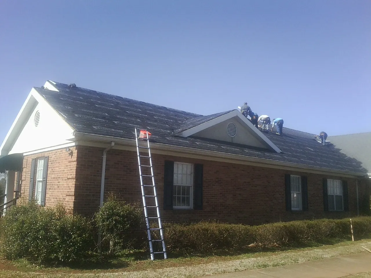 Crew tearing shingles off a brick ranch roof with a ladder set — Metal Roof Repair work by our crew in Saluda